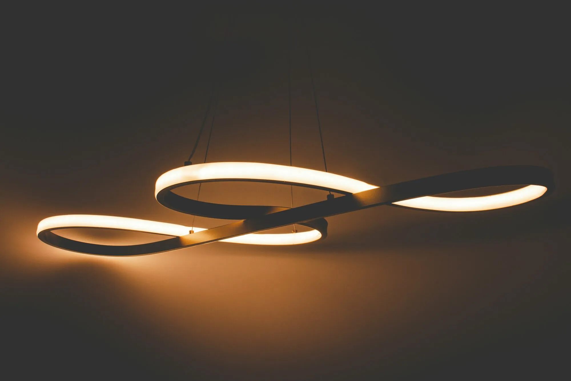 Gros plan sur une suspension LED aux courbes sculptées, allumée en blanc chaud sur fond sombre : la teinte basse de fin de soirée sur un luminaire choisi pour lui-même
