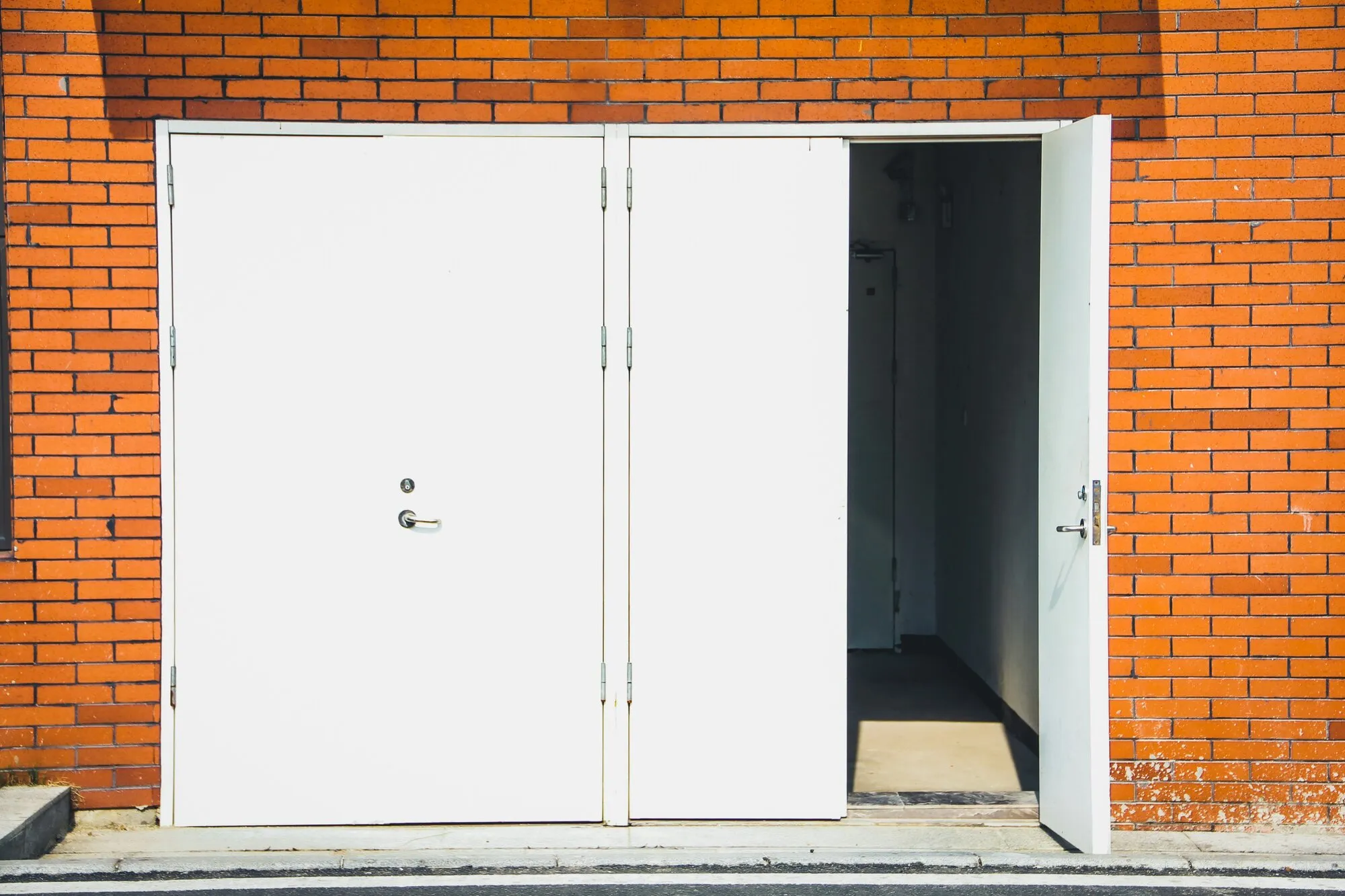 Rangée de portes blanches sur une façade en brique, toutes fermées sauf une : il suffit d’un seul accès laissé ouvert pour que l’intérieur soit visible depuis la rue